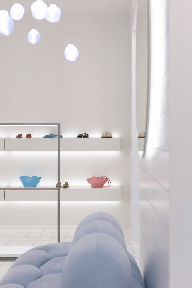 A minimalist store interior features a pastel blue sofa alongside wall shelves that display shoes and handbags one by one, all under white lighting that creates a clean, modern ambiance.