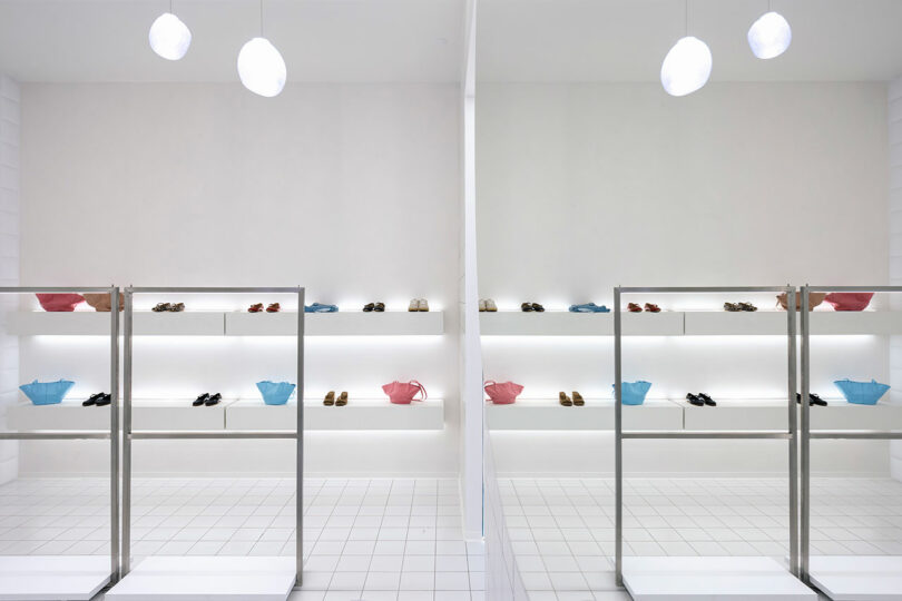 The minimalist shoe store interior, named "One by One," features white walls and bright lighting. Shoes and a few colorful baskets are elegantly displayed on pristine white shelves and in two sleek glass display cases.