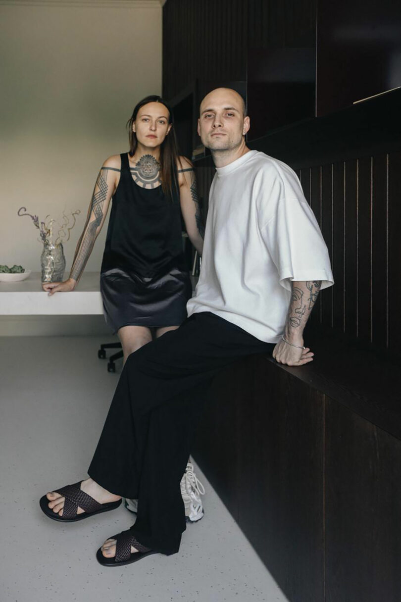 Two people with tattoos pose indoors; one is standing while the other is sitting on a bench.