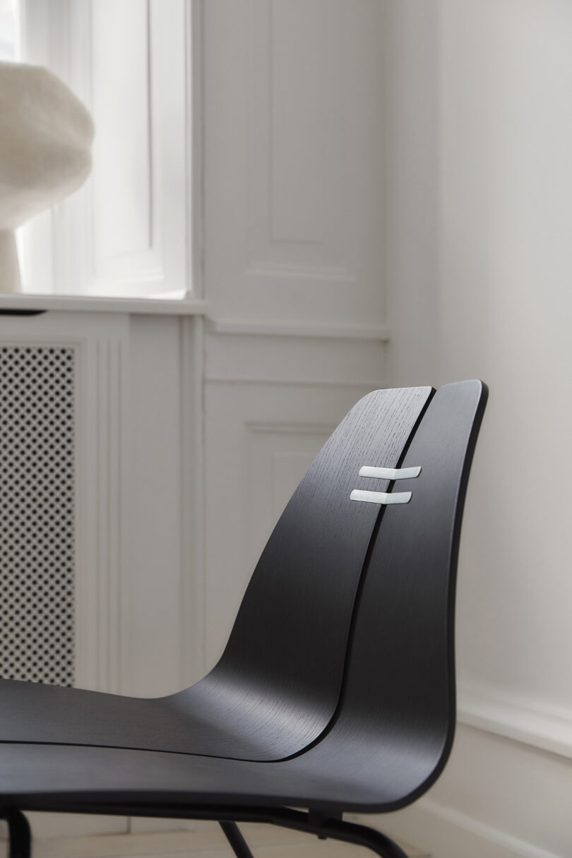 A modern black chair with a curved design is placed near a white wall with a radiator and a window in a bright room.