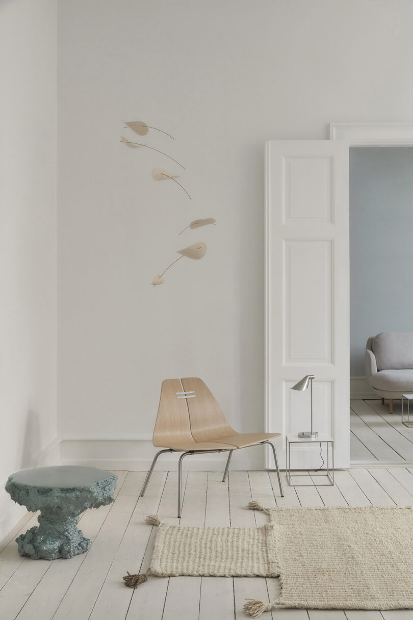 Minimalist room with a wooden chair, a textured rug, and a green sculptural side table. A hanging decorative piece is above the chair, and a gray sofa is visible through an open doorway.