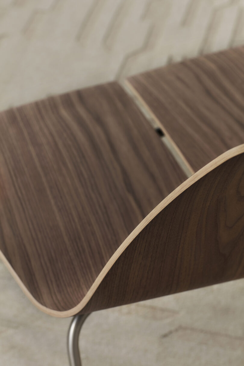 Close-up of a curved wooden chair with a visible seam in the middle. The chair has a natural wood grain finish and is set against a light, textured background.