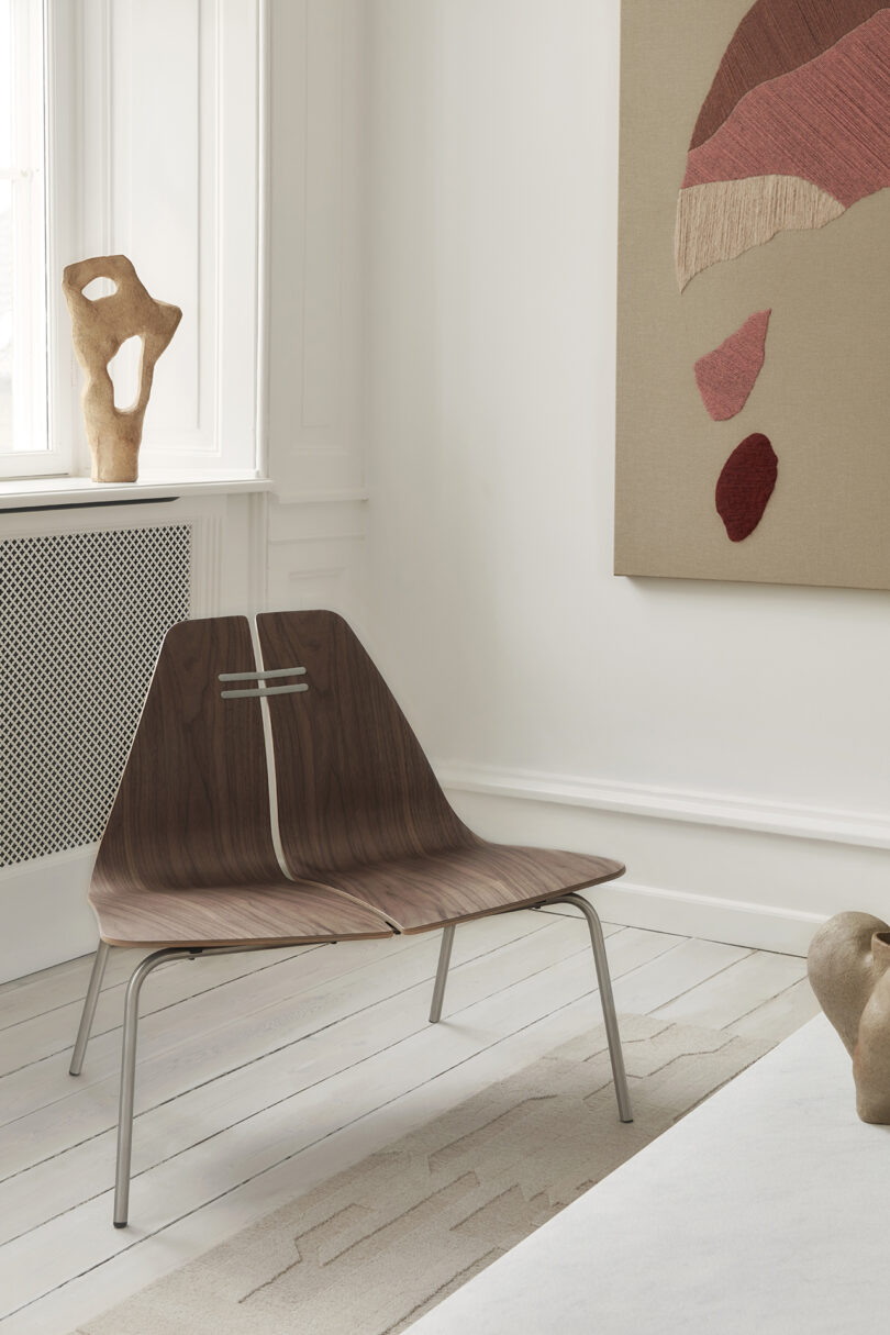 A modern wooden chair with metal legs sits in a minimalistic room with wooden flooring and abstract wall art. A sculptural piece decorates the windowsill.