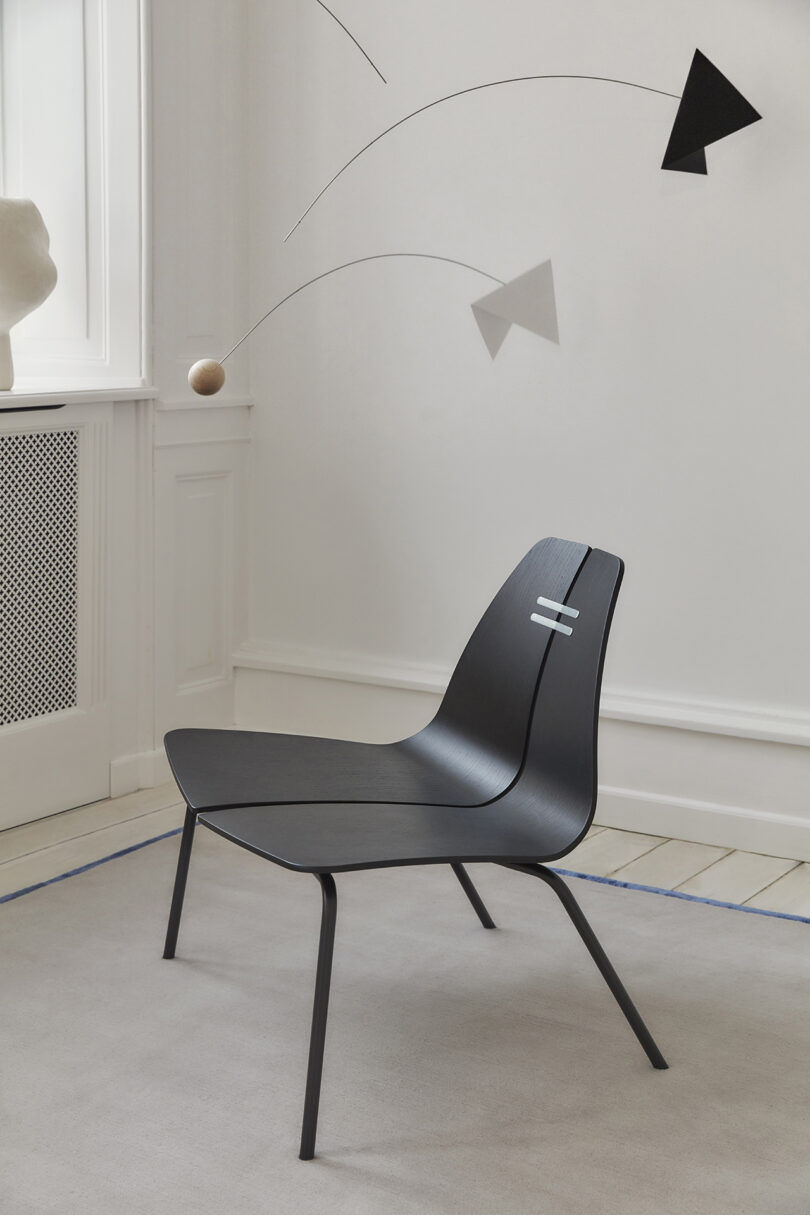 A modern, minimalist black chair with a curved seat and thin legs placed on a light gray rug. A geometric mobile hangs in the background near a window.