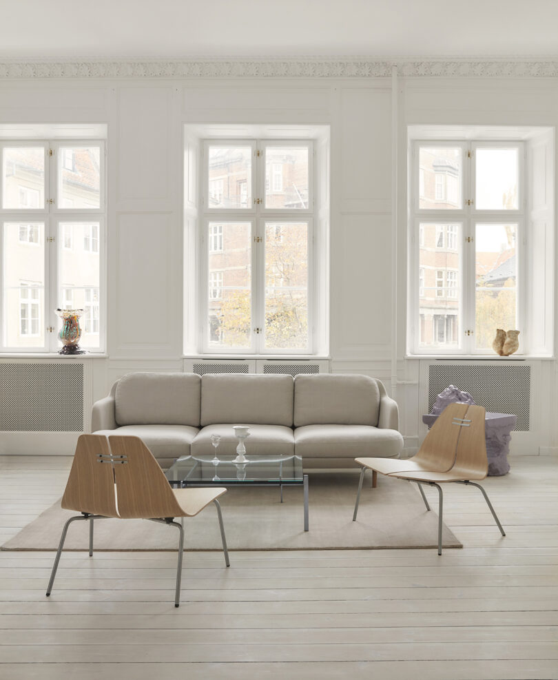 A minimalist living room with a beige sofa, glass coffee table, two wooden chairs, and large windows allowing natural light. A vase and small sculpture decorate the space.