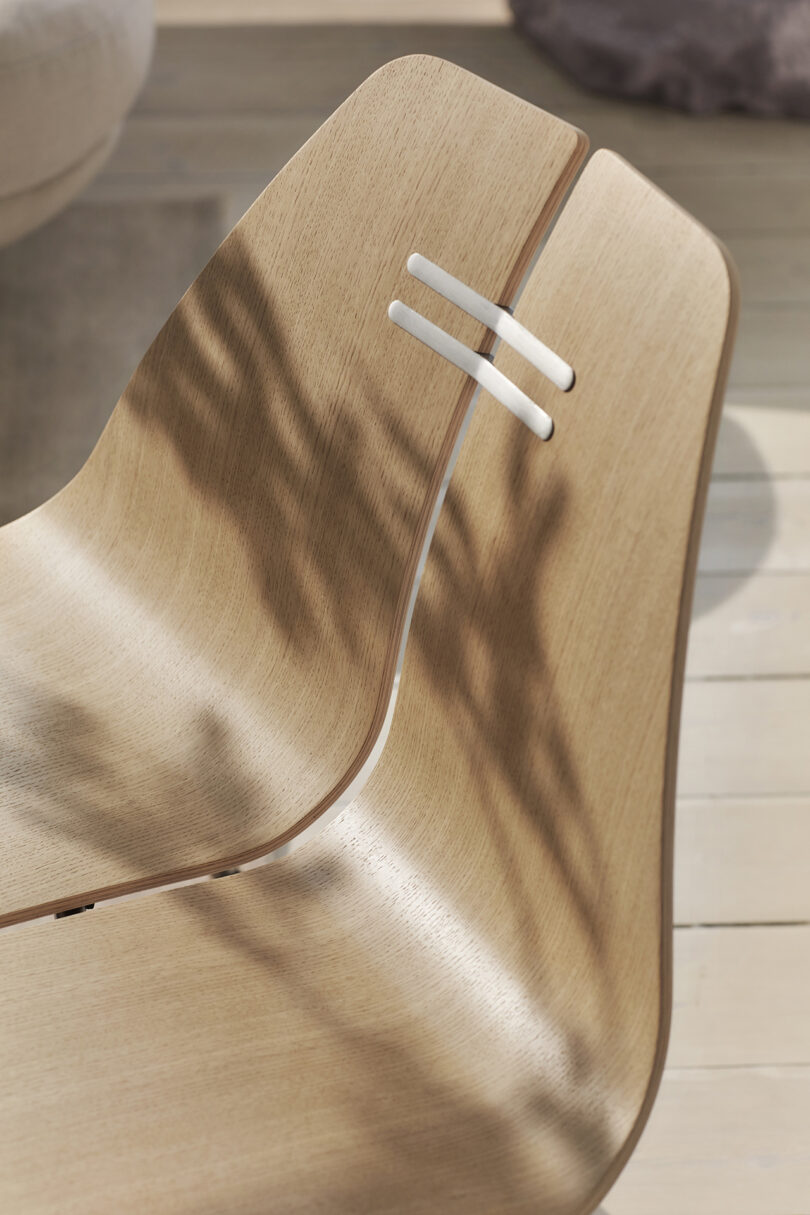 Close-up of a light wood chair with two white horizontal bars at the backrest, casting shadows. The seat and backrest form a continuous curve, with a smooth surface.