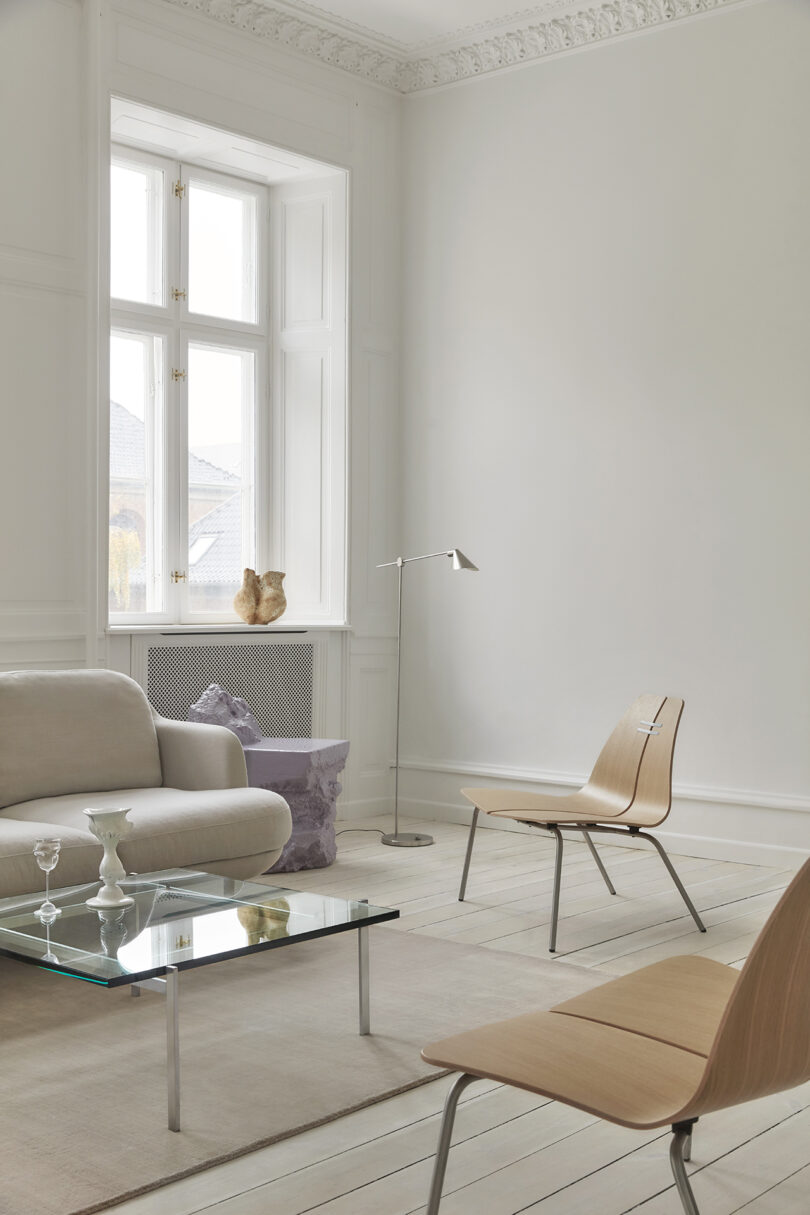 Minimalist living room with light wood furniture, glass coffee table, and a gray sofa. Large window with decorative molding and a small lamp. Neutral tones throughout.