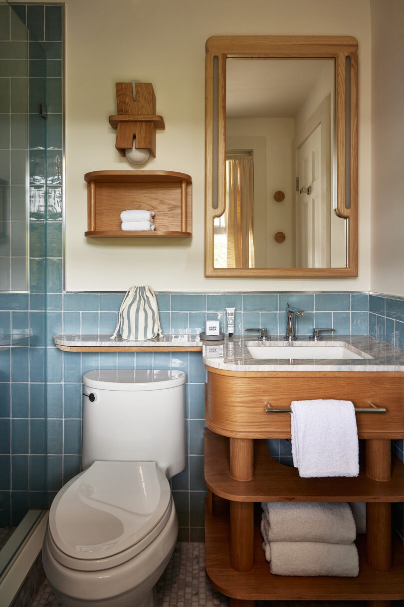 The bathroom, designed by Parts and Labor Design, features elegant blue-tiled walls, a toilet, and a wooden vanity with a sink and mirror. A striped accent pillow adds charm to the space, which also includes shelves neatly holding a folded towel and toiletries.