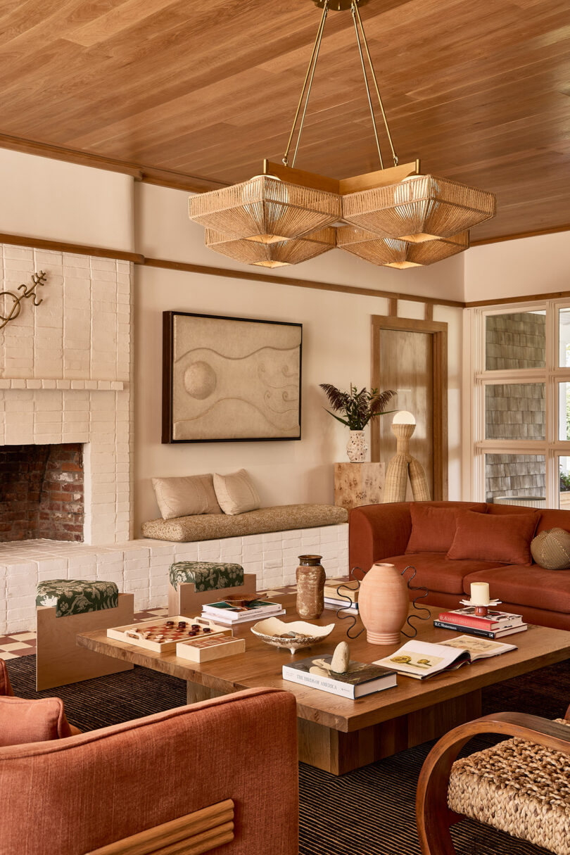 A cozy living room, inspired by Parts and Labor Design, features a brick fireplace, wooden ceiling, vibrant red sofas, and a large coffee table adorned with books and decor. Geometric pendant lights hang above while art and plants breathe life into the harmonious space.