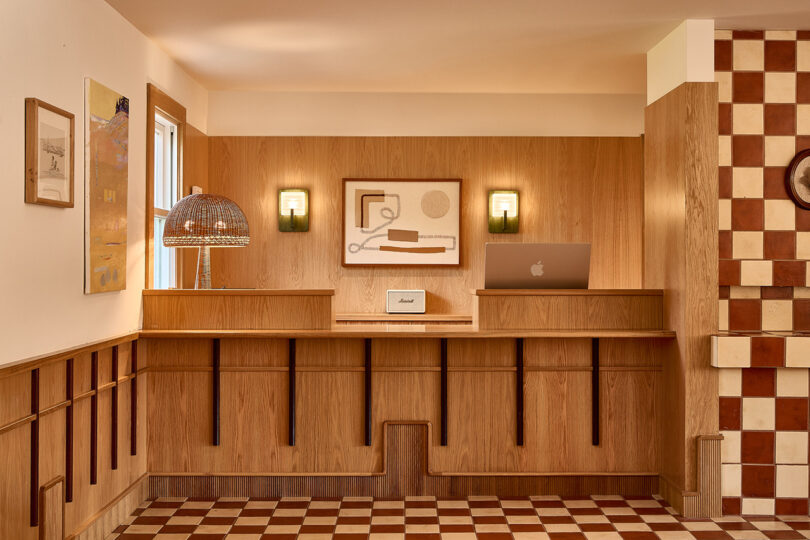 A cozy wooden reception desk by Parts and Labor Design features decorative art pieces, a lit lamp, and a laptop in a warmly lit room with checkered flooring and wood-paneled walls.