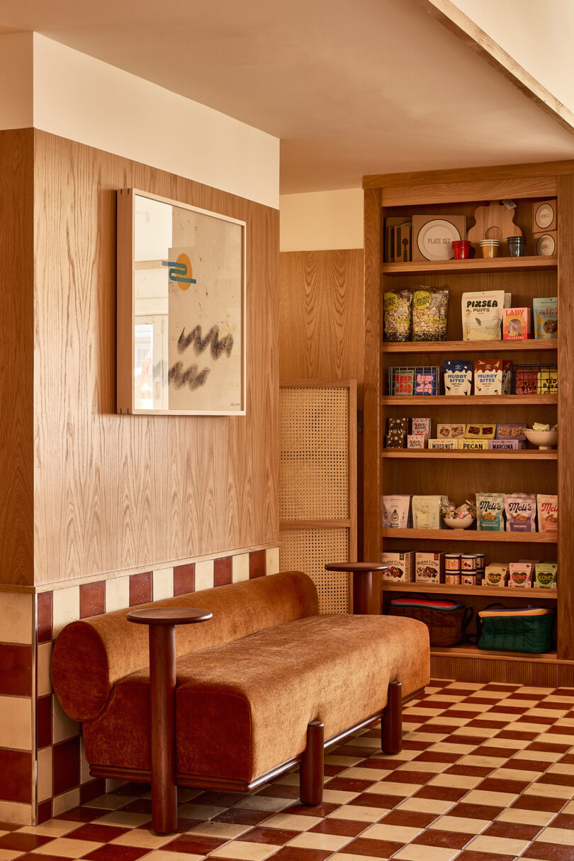 A cozy seating area featuring a brown bench and wooden panels, complemented by wall-mounted artwork, captures the essence of Parts and Labor Design. Shelves artfully display snacks and items against a stylish checkered brown and cream floor.