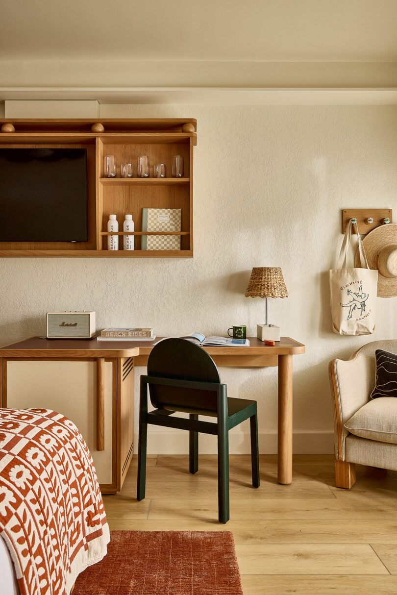 A hotel room desk with a black chair and wooden shelving crafted by Parts and Labor Design holds glasses, bottles, and a book. A tote bag hangs on the wall, while a bed with a patterned blanket is partially visible.