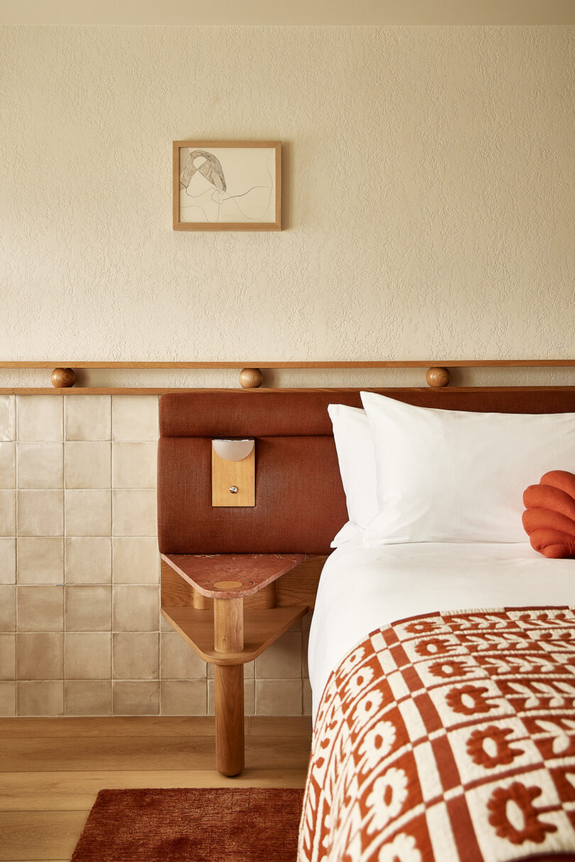 A cozy bedroom designed by Parts and Labor Design showcases a bed with a floral orange and white blanket, complemented by a stylish headboard and small table. A framed line drawing adds charm above the tan tiles and wood accents.