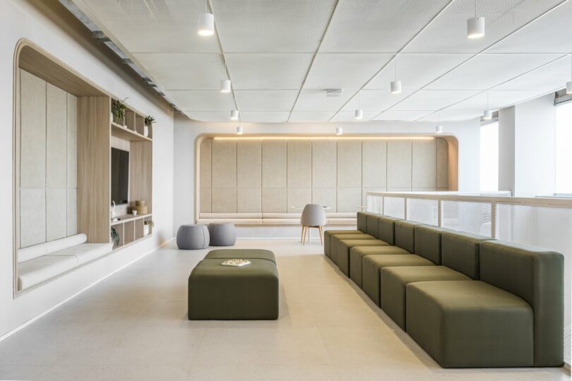 The Moas Group presents a modern lounge area featuring green modular seating and a central rectangular ottoman. Wall-integrated shelving and soft lighting enhance the ambiance, while light gray flooring and cream ceiling panels complete the sophisticated look.