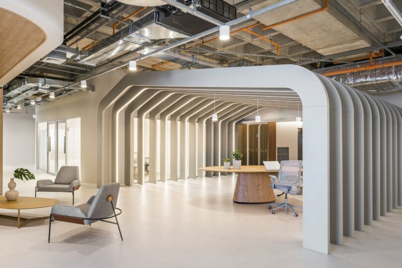 Modern office space featuring a geometric ceiling design, armchairs, a round wooden table, and a desk chair. Exposed ducts and stylish lighting add an industrial touch above.