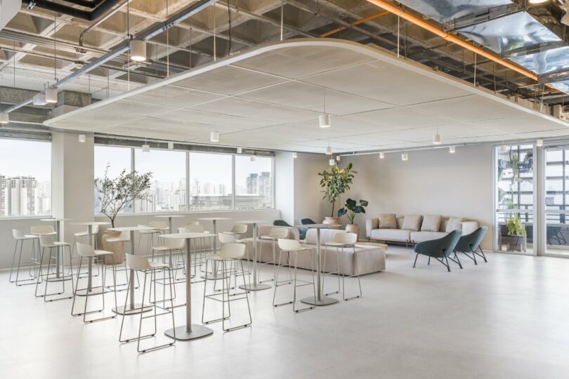 Moas Group's modern office lounge features white bar stools, tables, sofas, and plants. Large windows offer a stunning city view, while the exposed ceiling and bright lighting create an open atmosphere.