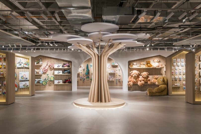 The modern store interior features a central tree-like display surrounded by shelves of toys and plush animals. Bright lighting and an industrial ceiling create a clean and open atmosphere.