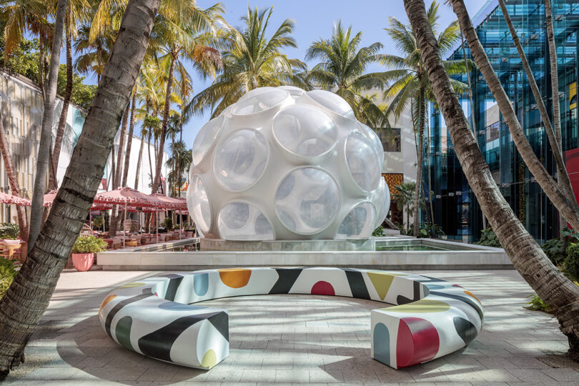 A white geodesic dome structure surrounded by palm trees and colorful benches in an outdoor plaza.