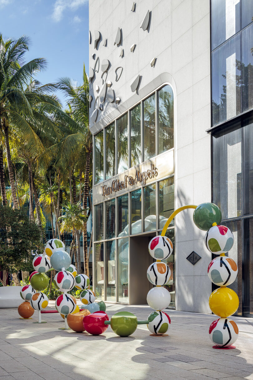 Colorful spherical sculptures are displayed outside a modern building with palm trees nearby.