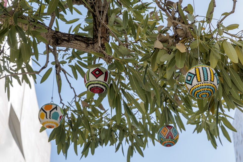 Tree branches with green leaves have several colorful beaded decorations hanging from them against a blue sky backdrop.