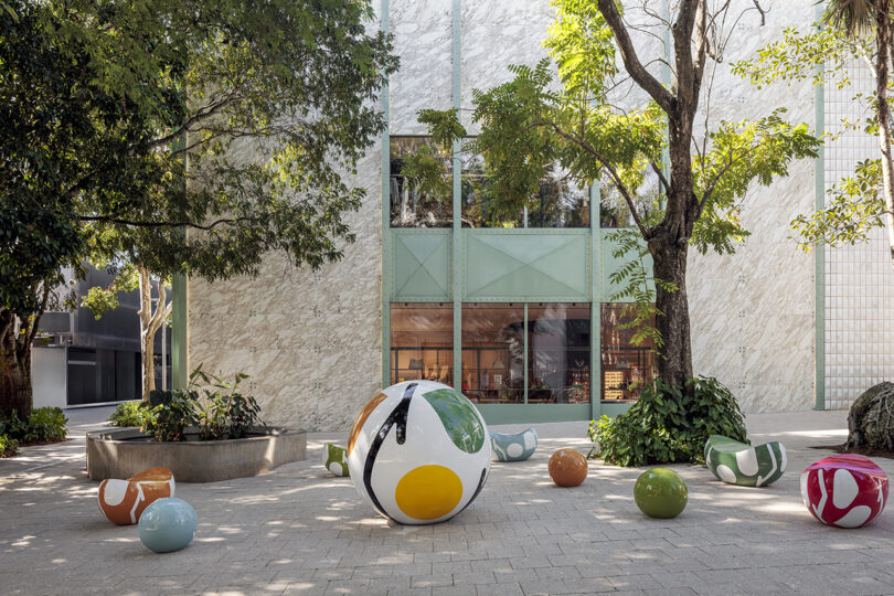 Colorful spherical sculptures are arranged in a courtyard with trees. A modern building with large windows stands in the background.