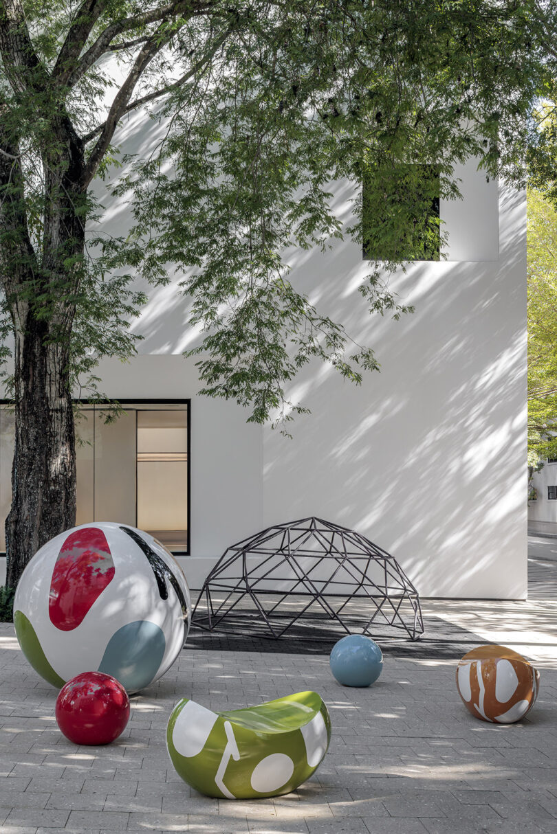 Outdoor art installation with colorful, round sculptures and a geometric metal structure in front of a white building with trees.