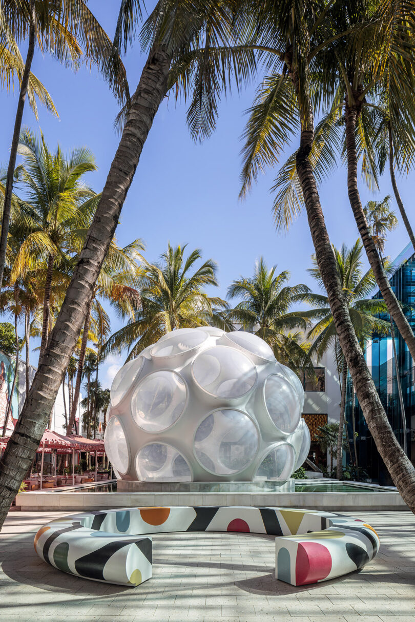 White geometric sculpture surrounded by palm trees and benches, with clear blue sky above.