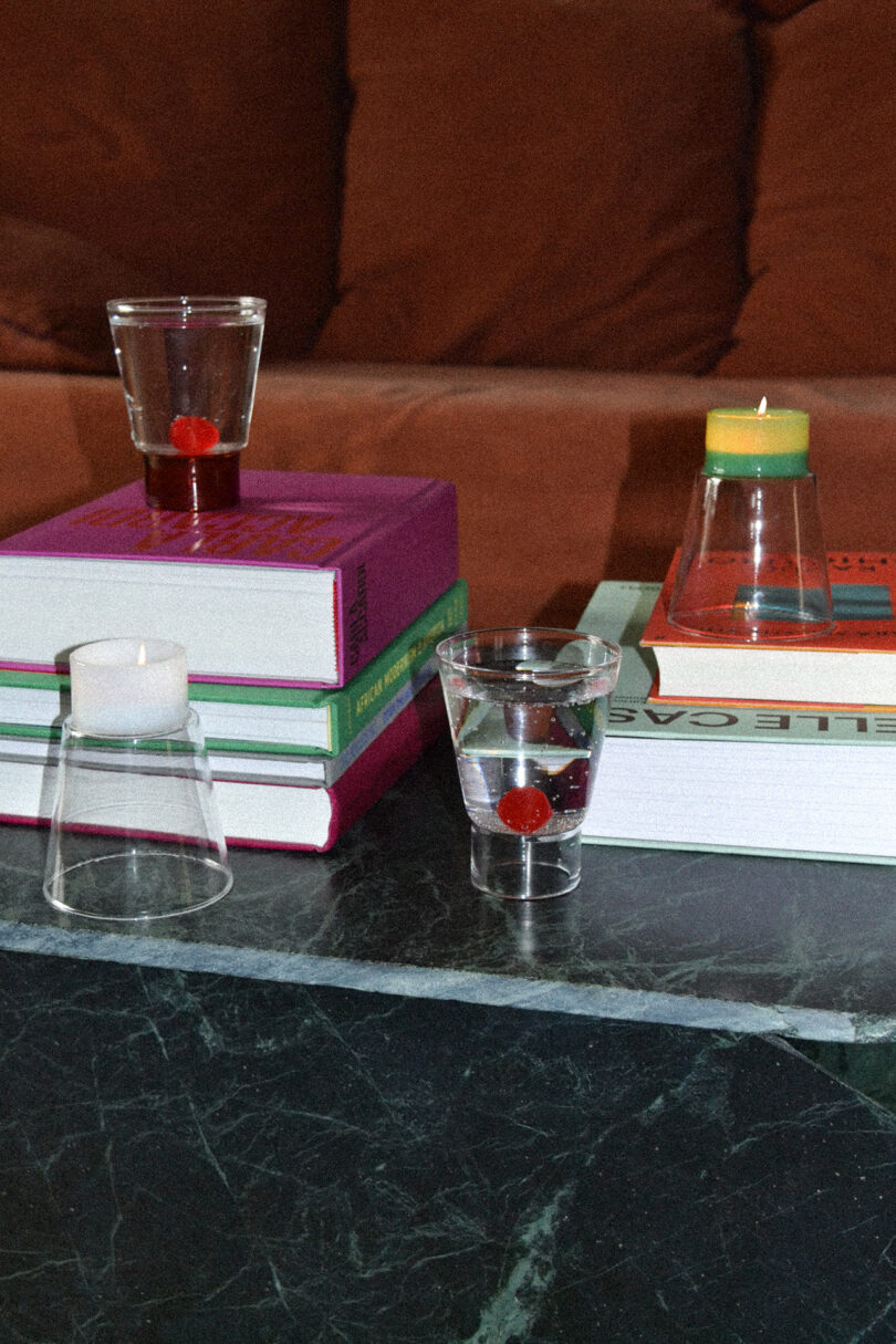 Two clear glasses with red liquid and a cherry sit on a stack of books and a marble surface, with an orange couch in the background.