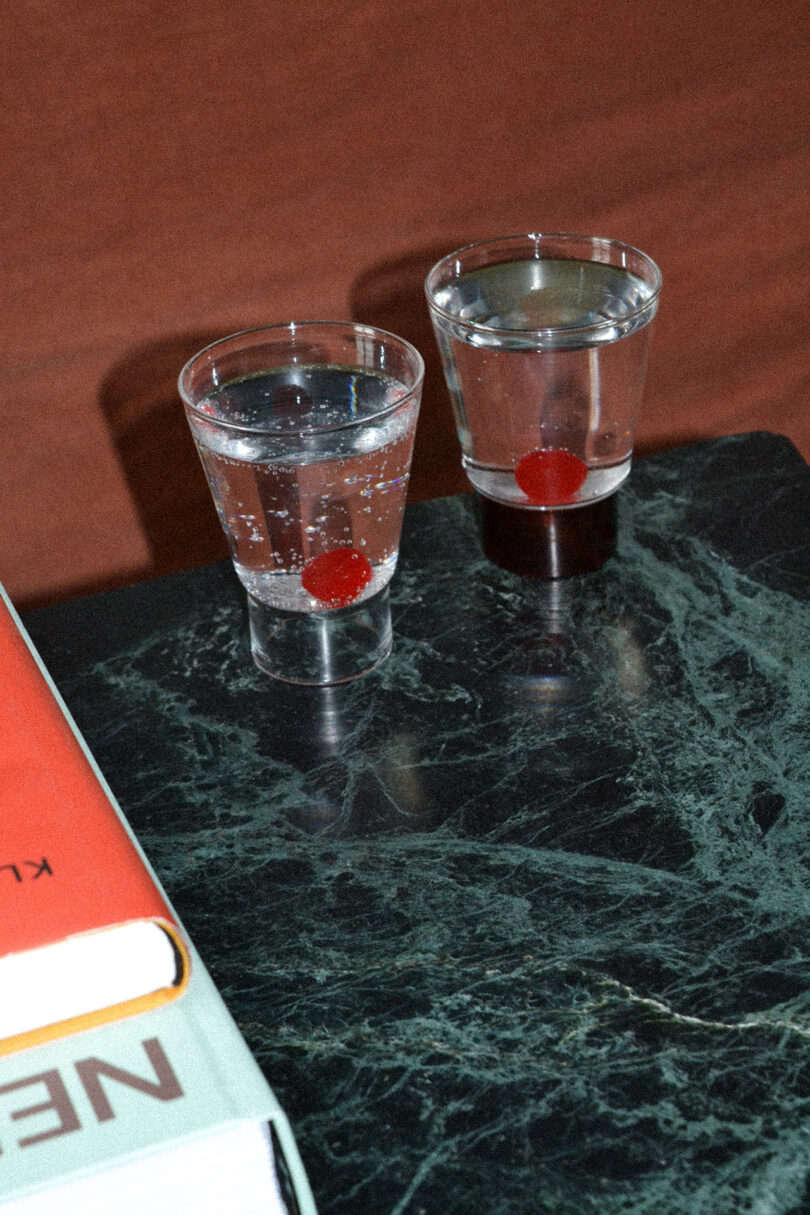 Two clear glasses with water and red cherries inside, placed on a dark green marble surface. Books partially visible at the edge.