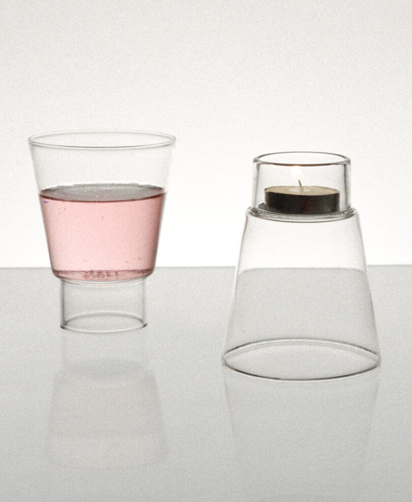 Two glass objects on a reflective surface: a cup with pink liquid on the left and an inverted cup with a small candle on top on the right.