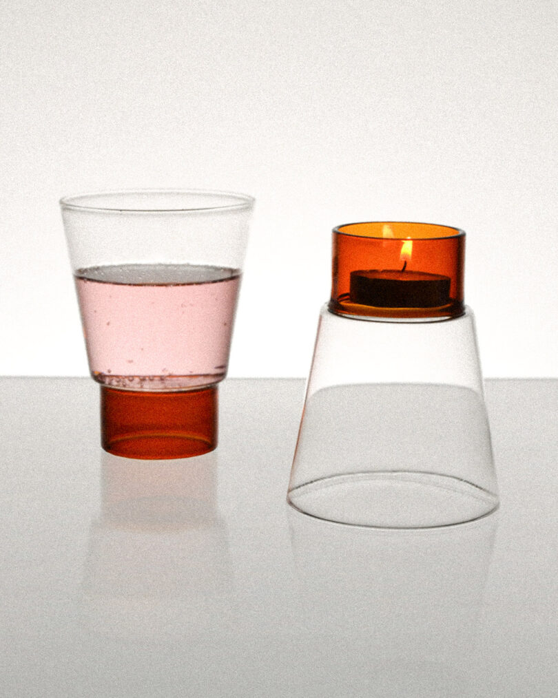 A pink drink in an orange glass next to an orange candle holder with a lit candle, all placed on a reflective surface.