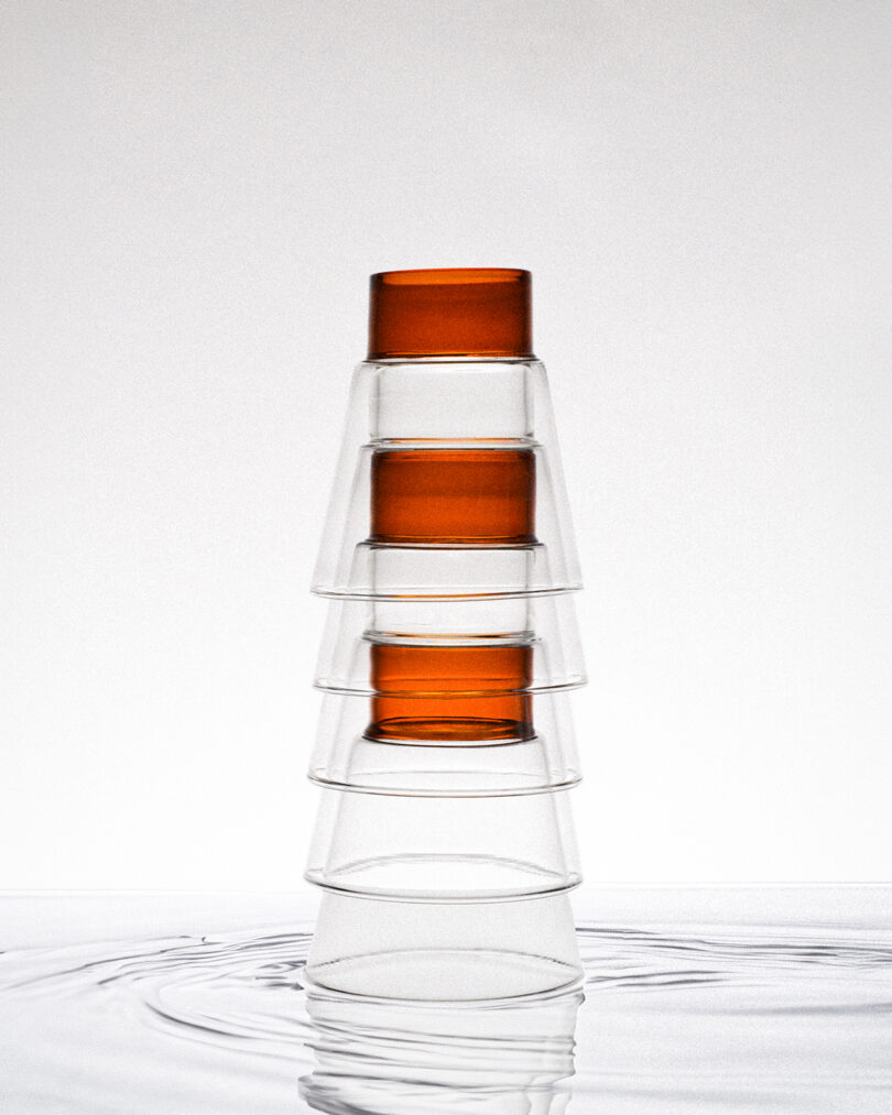 A stack of transparent glass cups with an orange cup on top, placed on a reflective surface.