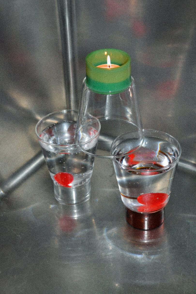 Two clear plastic cups with water and red Betta fish, next to an upside-down clear cup with a lit green candle on top, placed on a reflective metal surface.