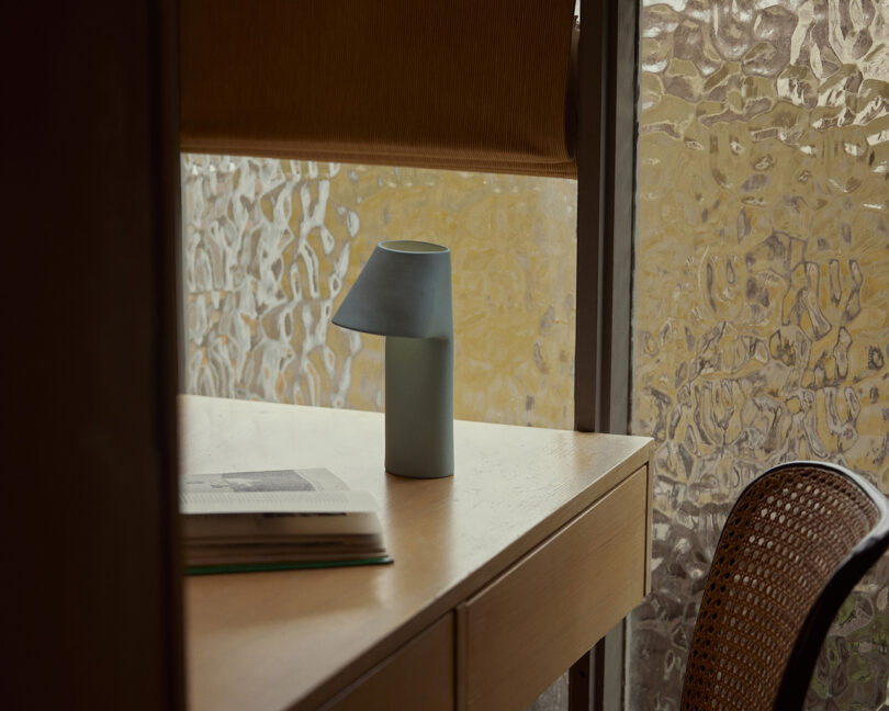 A small lamp on a wooden desk beside an open book. The desk is near a window with textured glass.