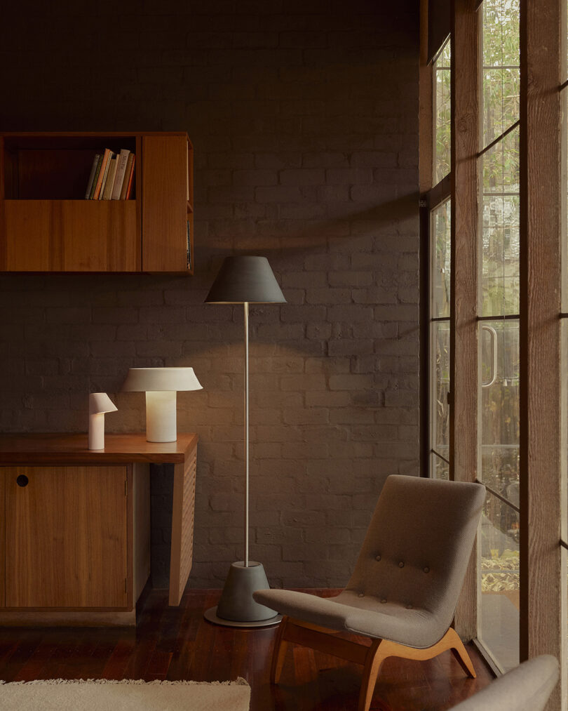 A cozy room with a gray chair, wooden desk, and two modern lamps. Books are placed on a wall-mounted shelf, and large windows allow natural light in.