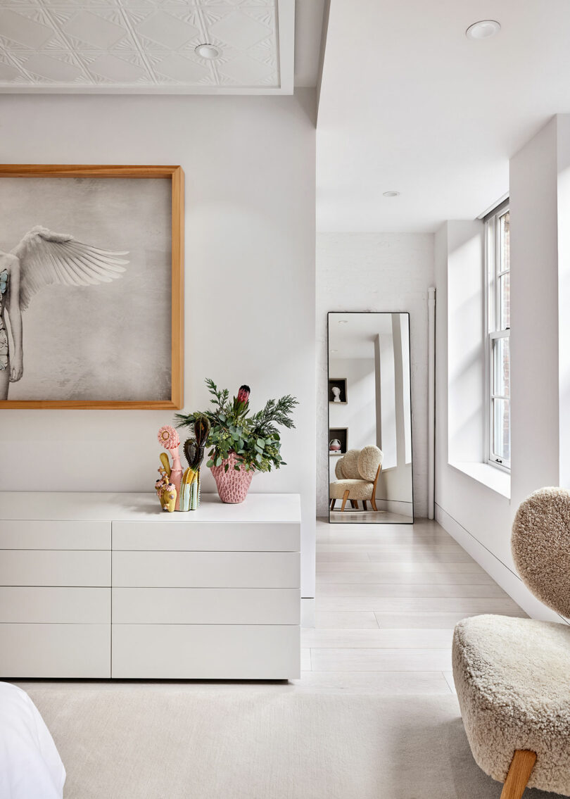 A modern bedroom with a white dresser, abstract art, and a potted plant. A tall mirror and a textured chair are near the window, reflecting natural light.