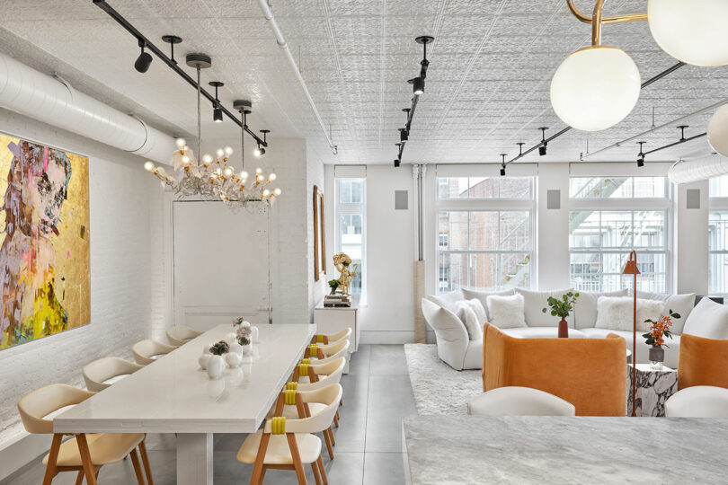 Modern open-plan living space with a large dining table, white seating, abstract art on the wall, and spherical lights. Large windows provide natural light.