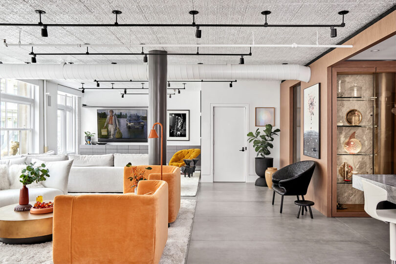 Modern living room with orange chairs, a white sofa, artwork, a marble table, plants, and track lighting. A glass display cabinet is on the right.