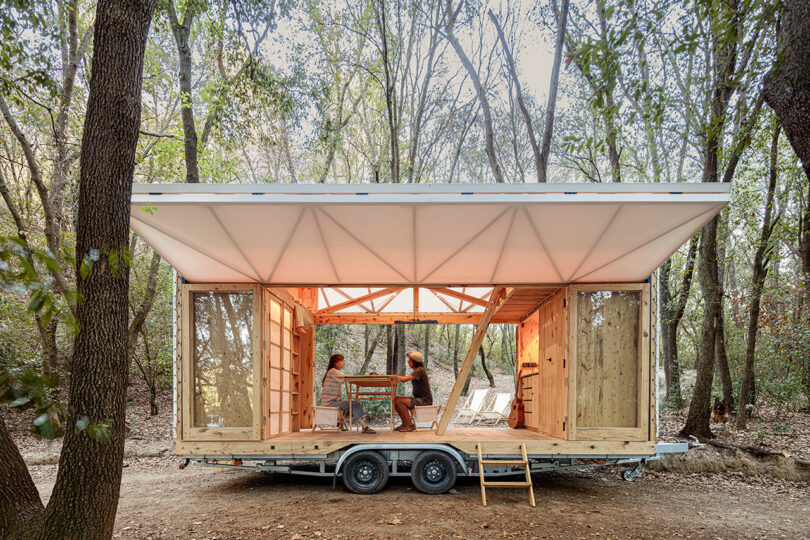 A small, mobile wooden cabin with open sides is situated in a forest. Two people are seated inside at a table, engaged in conversation.