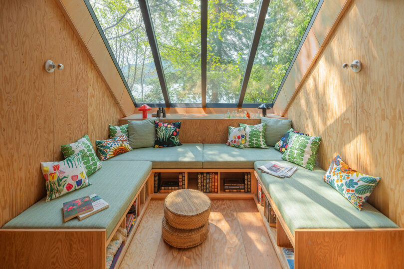 Cozy corner with a skylight, featuring a built-in wooden bench with cushions and colorful pillows, surrounded by books and wicker stools. Lush greenery visible through the window.