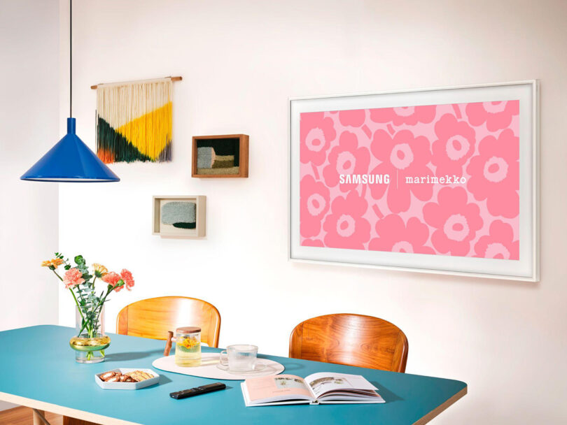 A modern dining area with a teal table, wooden chairs, a blue pendant lamp, and a Samsung Marimekko art frame on the wall. Wall decorations and a vase with flowers are also visible.