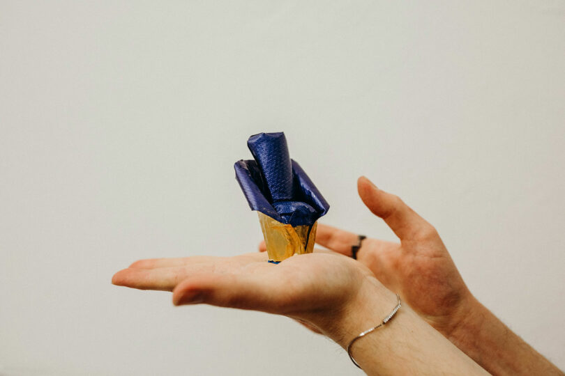 Hands holding a small cone wrapped in gold and blue paper against a plain background.