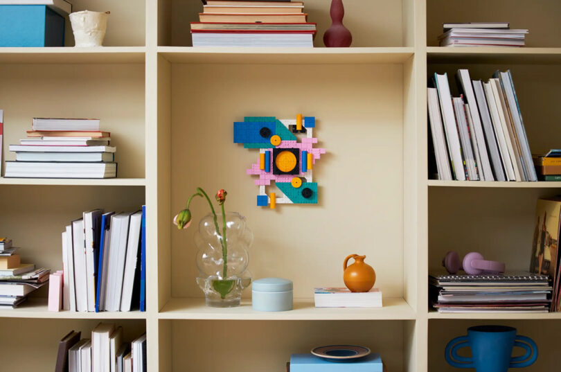 Wall-mounted bookshelf with various books, a geometric artwork, and decorative items, including a glass vase with drooping flowers and a small orange pitcher.