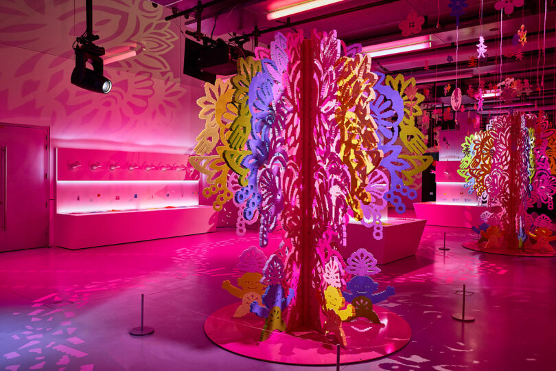 A brightly lit room features large, colorful cut-out paper art installations, with intricate designs and pink lighting.