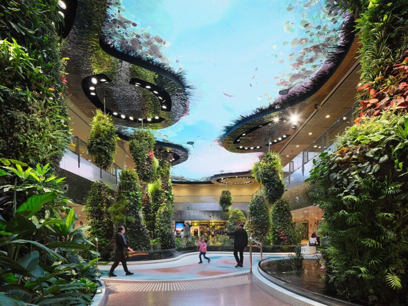 Modern atrium with tall vertical gardens and a ceiling display featuring aquatic imagery. Visitors walk through the bright, green-filled space