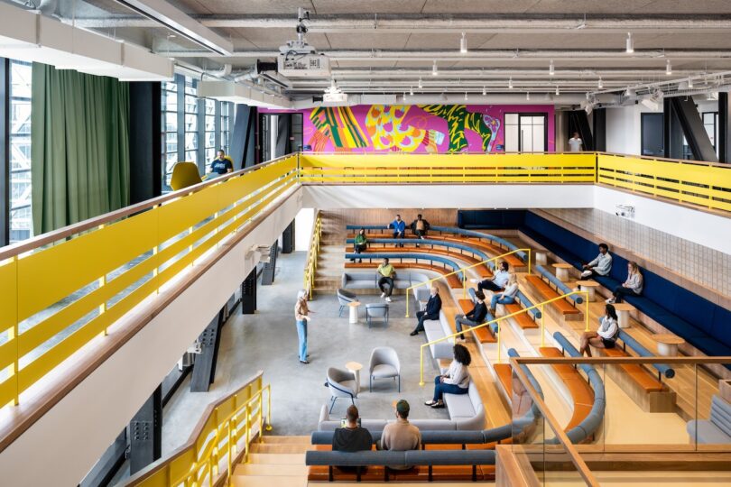 A modern, open two-story workspace with people seated on tiered wooden benches. A colorful mural decorates the upper wall