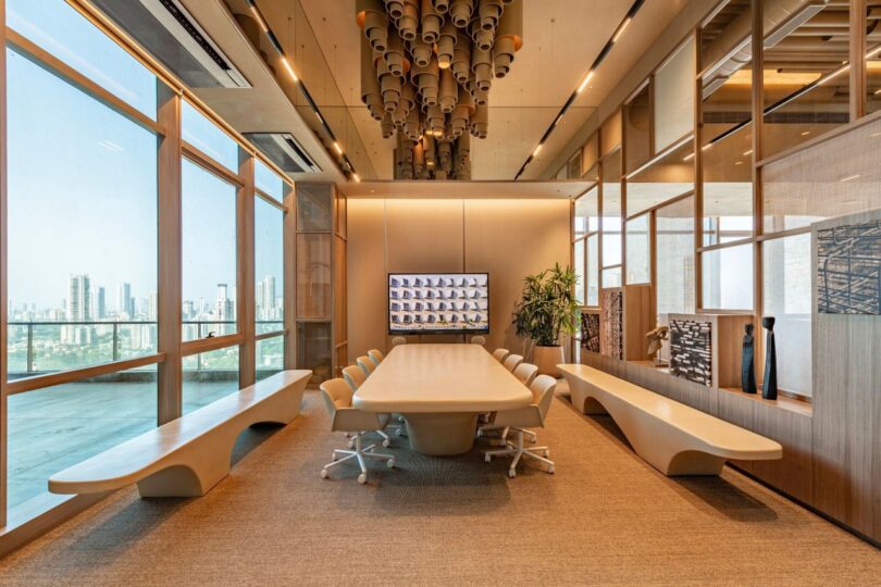 A modern conference room with a large table, swivel chairs, and a video screen on the wall. Floor-to-ceiling windows offer a cityscape view. Ceiling features artistic light fixtures