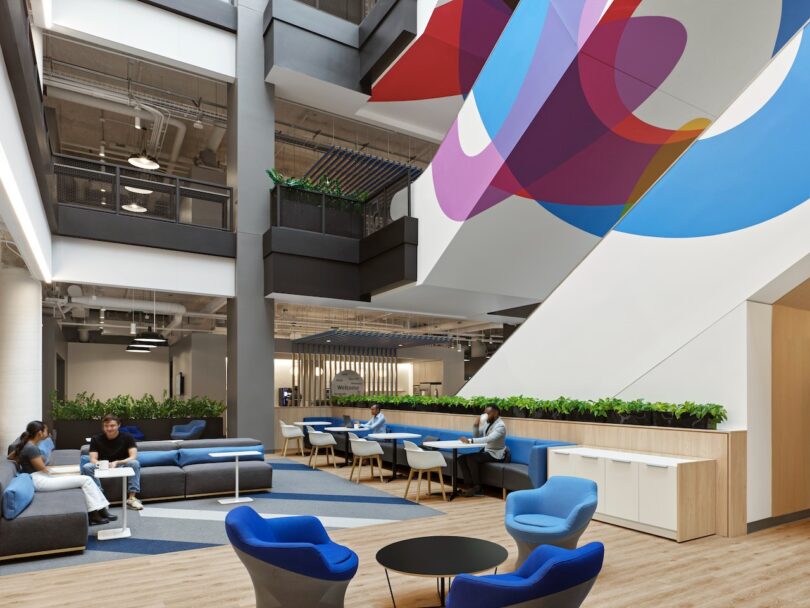 Modern office interior with colorful wall art, people sitting in blue chairs and on sofas, and tables with greenery