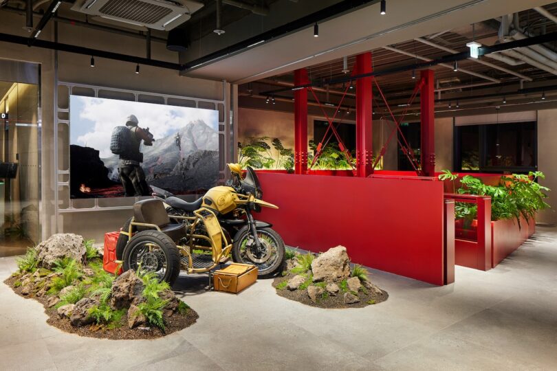 A themed room with a decorated motorcycle display, rocks, and plants. A large mural on the wall depicts a soldier in a mountainous setting. Red structures and potted plants are in the background