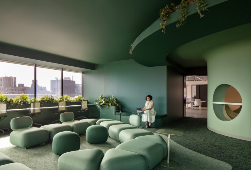 A person sits with a laptop in a modern, green-themed office lounge with round furniture and plants, overlooking a cityscape through large windows